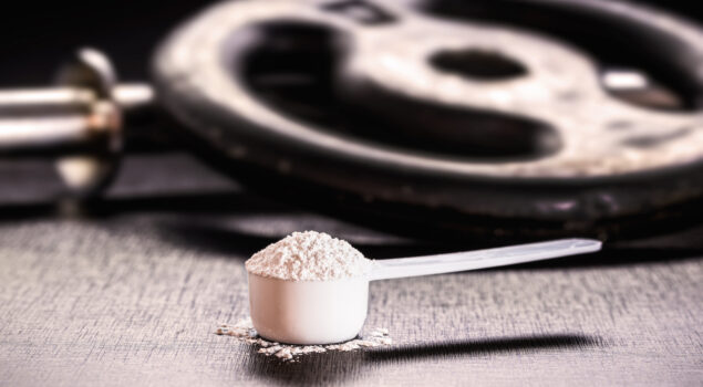 Imagem mostra um "scoop" cheio de creatina ao lado de anilhas de peso para exemplificar os benefícios da creatina no treino.